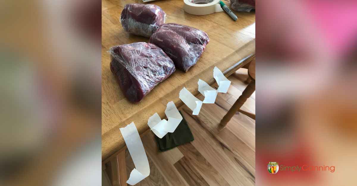 Pieces of masking tape hanging off the edge of the kitchen table with wrapped meat behind it.