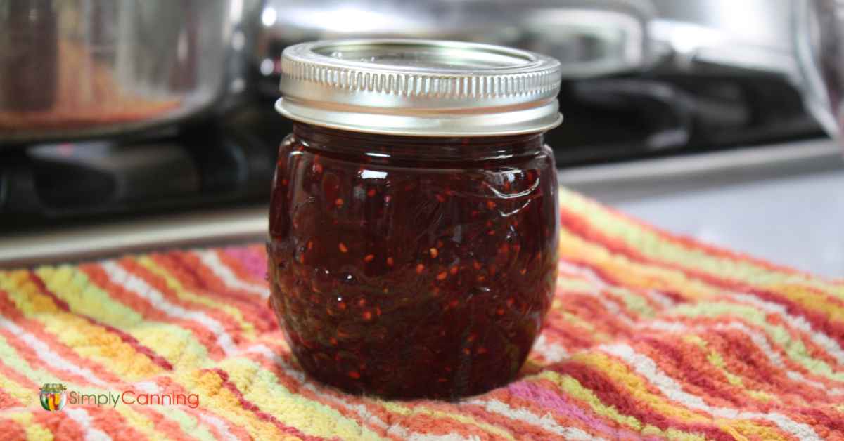 Pretty jam jar with strawberry raspberry jam sitting on a red, orange and yellow striped dishcloth.  